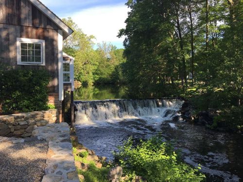 A serene scene featuring a small waterfall beside a house, surrounded by lush greenery and a calm river.