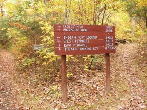 Wooden trail sign in a forest, indicating distances to various lookout points and a parking lot. Fall foliage surrounds it.