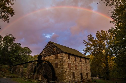 A historic stone mill with a large water wheel under a vibrant rainbow and cloudy sky.