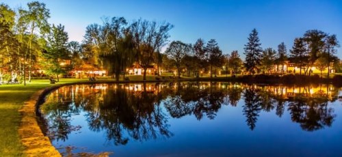 A serene park at dusk, featuring a calm pond reflecting trees and lights from nearby buildings.