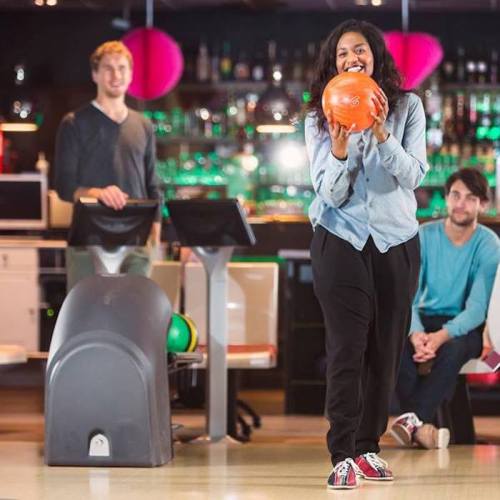A woman smiles while holding a bowling ball, with two men watching her in a bowling alley.