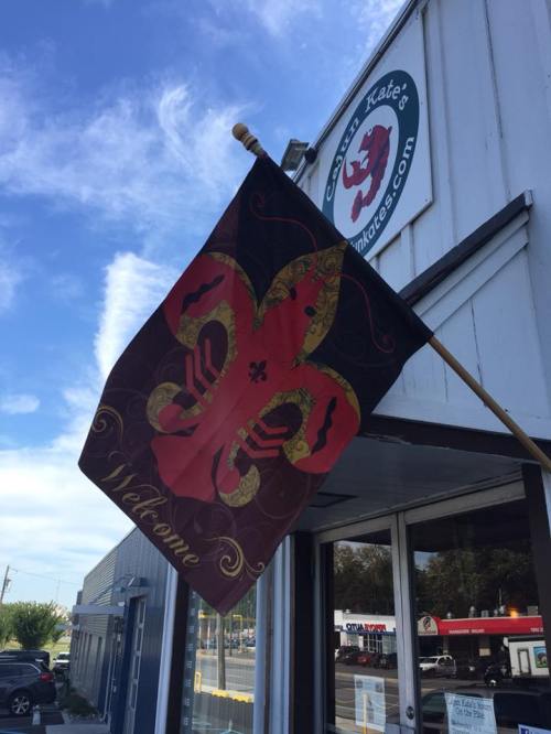 A colorful flag with a fleur-de-lis design and "Welcome" text, displayed outside a shop named "Cajun Kate's."