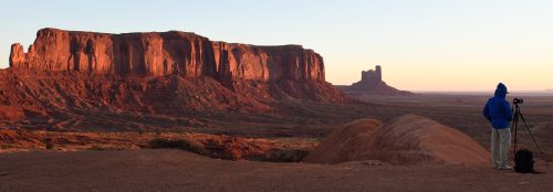 A photographer in a blue hoodie captures the sunrise over Monument Valley's red rock formations.