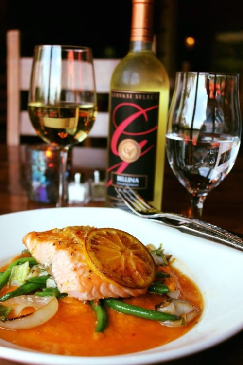 A plate of salmon with lemon, served on a bed of vegetables, accompanied by glasses of white wine and water.