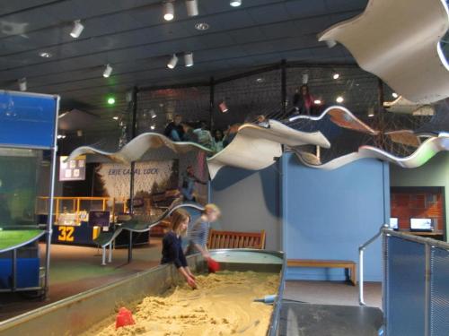 Children playing in a sand area at a museum, with a climbing structure and other kids above them.
