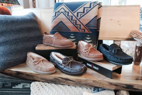 A display of various styles of cozy shoes on a wooden shelf, with a patterned blanket in the background.