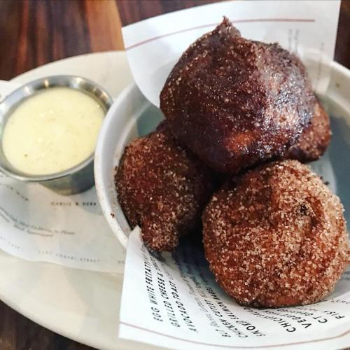 A bowl of golden-brown fried dough balls dusted with sugar, served with a small cup of creamy dipping sauce.