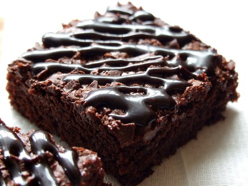 A close-up of a chocolate brownie drizzled with rich chocolate sauce on a light background.