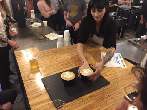 A barista serves two cups of latte art on a wooden table at a coffee event, with people in the background.