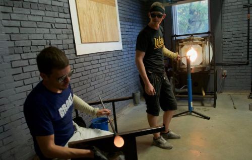 Two glassblowers work in a studio, one shaping glass on a rod while the other monitors a furnace with molten glass.