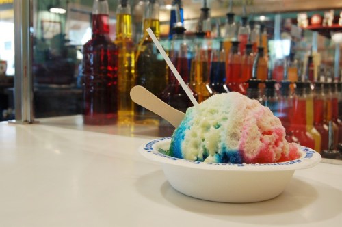 A colorful snow cone in a white bowl with a wooden stick, set against a backdrop of vibrant syrup bottles.