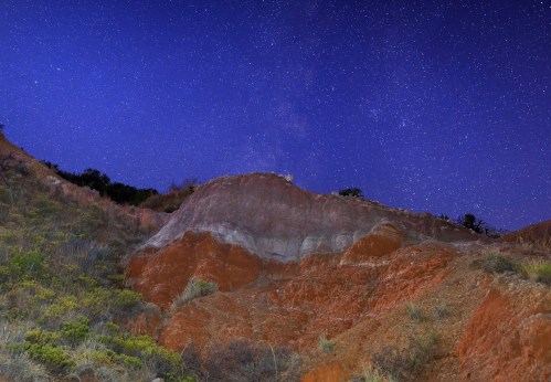 A rocky landscape under a starry night sky, featuring reddish earth and sparse vegetation.
