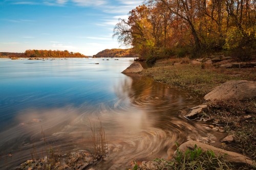 A serene river scene with swirling water, surrounded by autumn foliage and rocky banks under a clear blue sky.