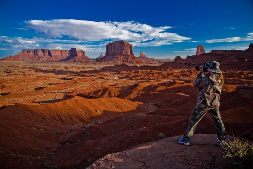 A person in camouflage takes a photo of a vast desert landscape with red rock formations and blue skies.