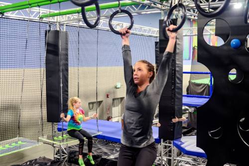 A woman performs a pull-up on gym rings while a girl plays on nearby equipment in a colorful indoor gym.