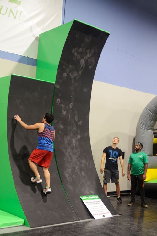 A person climbs a curved wall while two others watch in a gym setting with inflatable structures in the background.