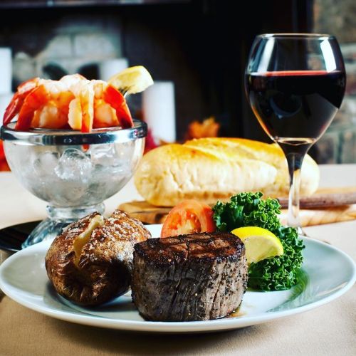 A plate with a grilled steak, baked potato, and salad, accompanied by shrimp cocktail, a glass of red wine, and bread.