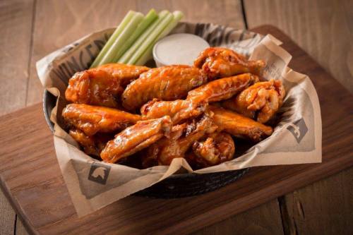 A basket of spicy chicken wings with celery sticks and a side of dipping sauce on a wooden table.