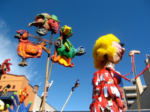 Colorful puppets and figures on sticks against a bright blue sky, showcasing vibrant art and creativity.