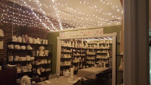 A cozy pottery studio with shelves of white ceramic pieces, warm lighting, and a table set for crafting.