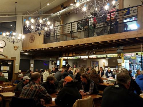 A bustling bar interior with patrons seated at tables, a two-level layout, and chandeliers overhead.
