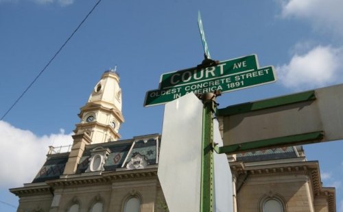 Street sign for Court Ave, noting it as the oldest concrete street in America, with a historic building in the background.
