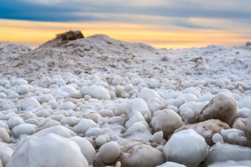 A close-up of icy, white formations on a beach at sunset, with soft colors in the sky and a sandy mound in the background.