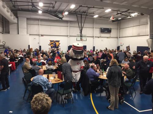 A crowded indoor event with people seated at tables, featuring a mascot in a baseball uniform among the attendees.