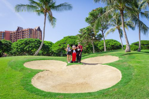 A group of three people poses in a large sand trap shaped like Mickey Mouse, surrounded by palm trees and greenery.
