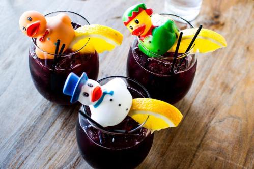 Three colorful cocktails with rubber duck and snowman decorations, garnished with lemon slices, on a wooden table.
