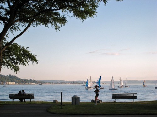A serene lakeside scene with sailboats on the water, joggers, and people sitting on benches under trees.
