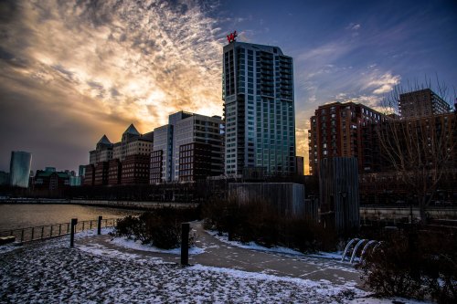 City skyline at sunset with modern buildings, a river, and a snowy foreground. Dramatic clouds fill the sky.