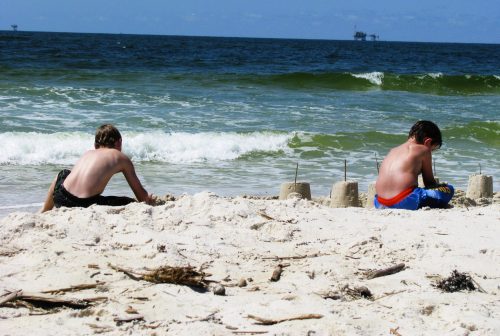 Two boys build sandcastles on a beach, with waves crashing nearby and an oil rig visible in the distance.