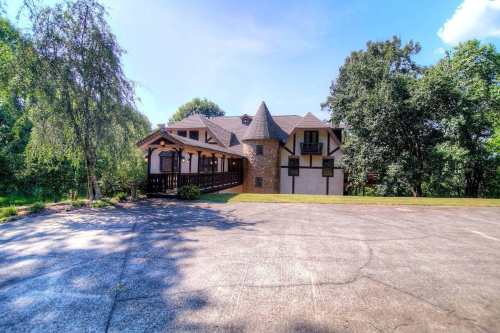 A large, unique house with a turret, surrounded by trees and a spacious driveway under a clear blue sky.