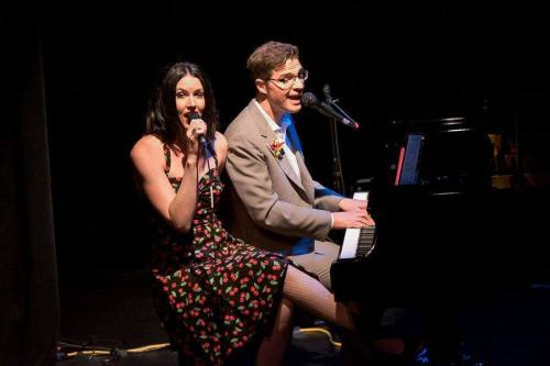 A singer in a floral dress performs alongside a pianist on stage, both engaged in a lively musical performance.
