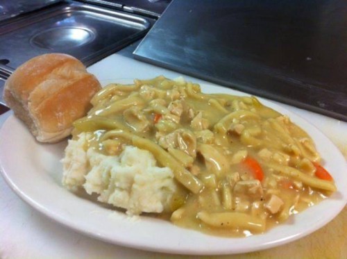 A plate of chicken and noodles with vegetables, served with mashed potatoes and a roll on the side.