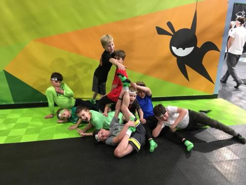 A group of children in colorful outfits playfully piled together on a green and black mat, smiling and having fun.