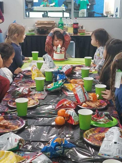 A child blows out candles on a birthday cake surrounded by friends at a festive table with snacks and drinks.