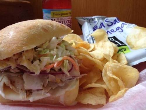 A sandwich with shredded chicken and coleslaw, served with potato chips and a bottle of pomegranate drink.