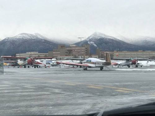 A snowy airport scene with several small planes parked, mountains in the background under a cloudy sky.