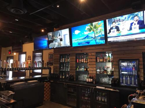 Interior of a bar featuring a wooden wall, multiple TVs, a variety of drinks, and a tap system.
