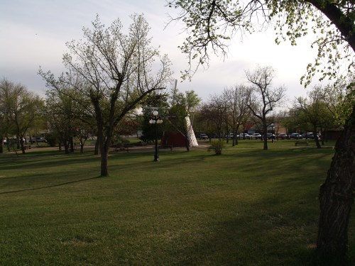 A park scene with green grass, trees, and a teepee in the background under a cloudy sky.