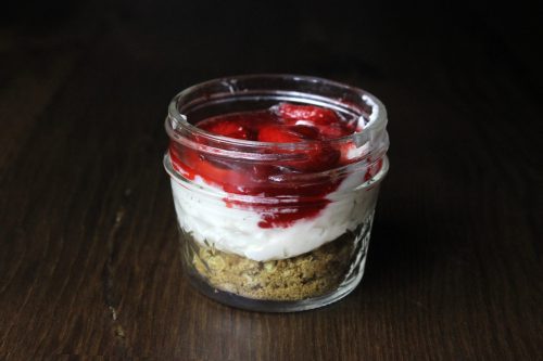 A jar filled with layers of graham cracker crust, creamy yogurt, and strawberry topping.