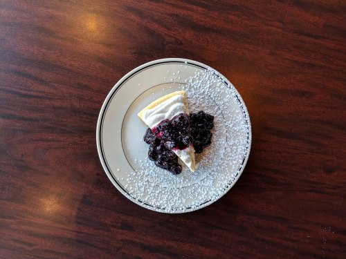 A slice of cheesecake topped with blueberry compote and dusted with powdered sugar on a white plate.