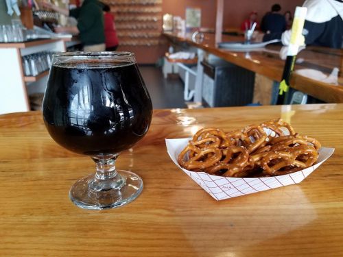 A dark beer in a glass next to a serving of pretzels on a wooden bar counter.