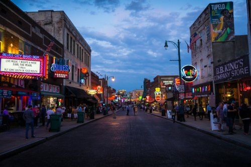 A lively street scene at dusk, featuring neon signs, music venues, and people enjoying the atmosphere in a vibrant city.