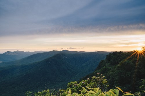 A scenic view of mountains under a cloudy sky, with the sun setting on the horizon and lush greenery in the foreground.