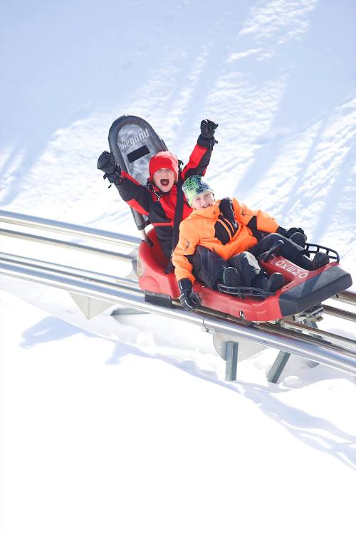 Two children joyfully ride a sled down a snowy track, with bright winter clothing and smiles on their faces.