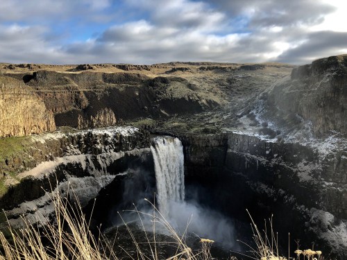 A stunning waterfall cascades into a rocky canyon, surrounded by rugged cliffs and grassy terrain under a cloudy sky.