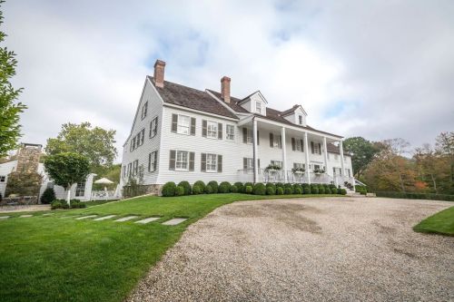 A large white colonial-style house with a gravel driveway and manicured lawns, surrounded by trees under a cloudy sky.